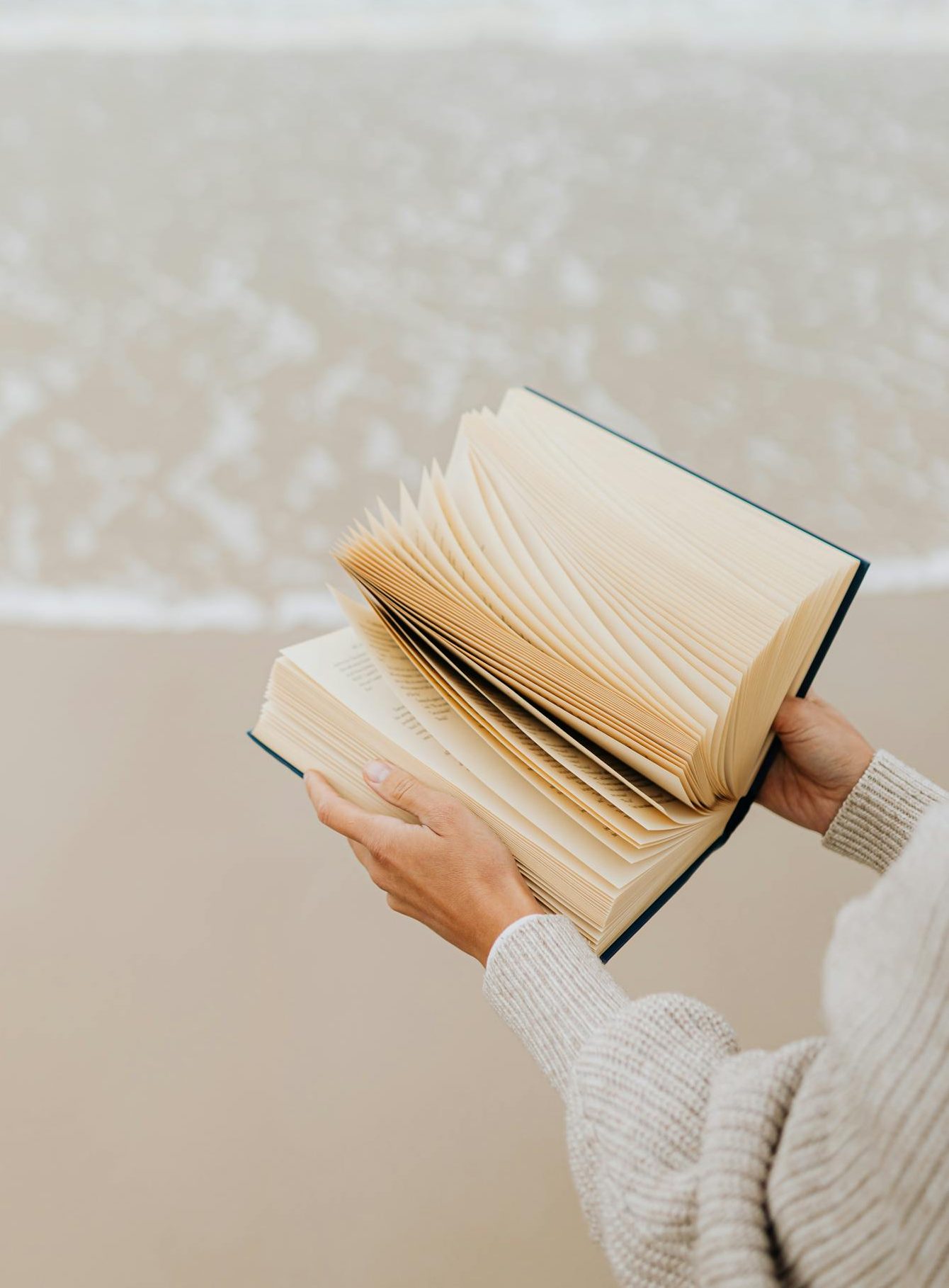 Hands holding a book with flipping pages by the seaside, capturing a peaceful beach moment.